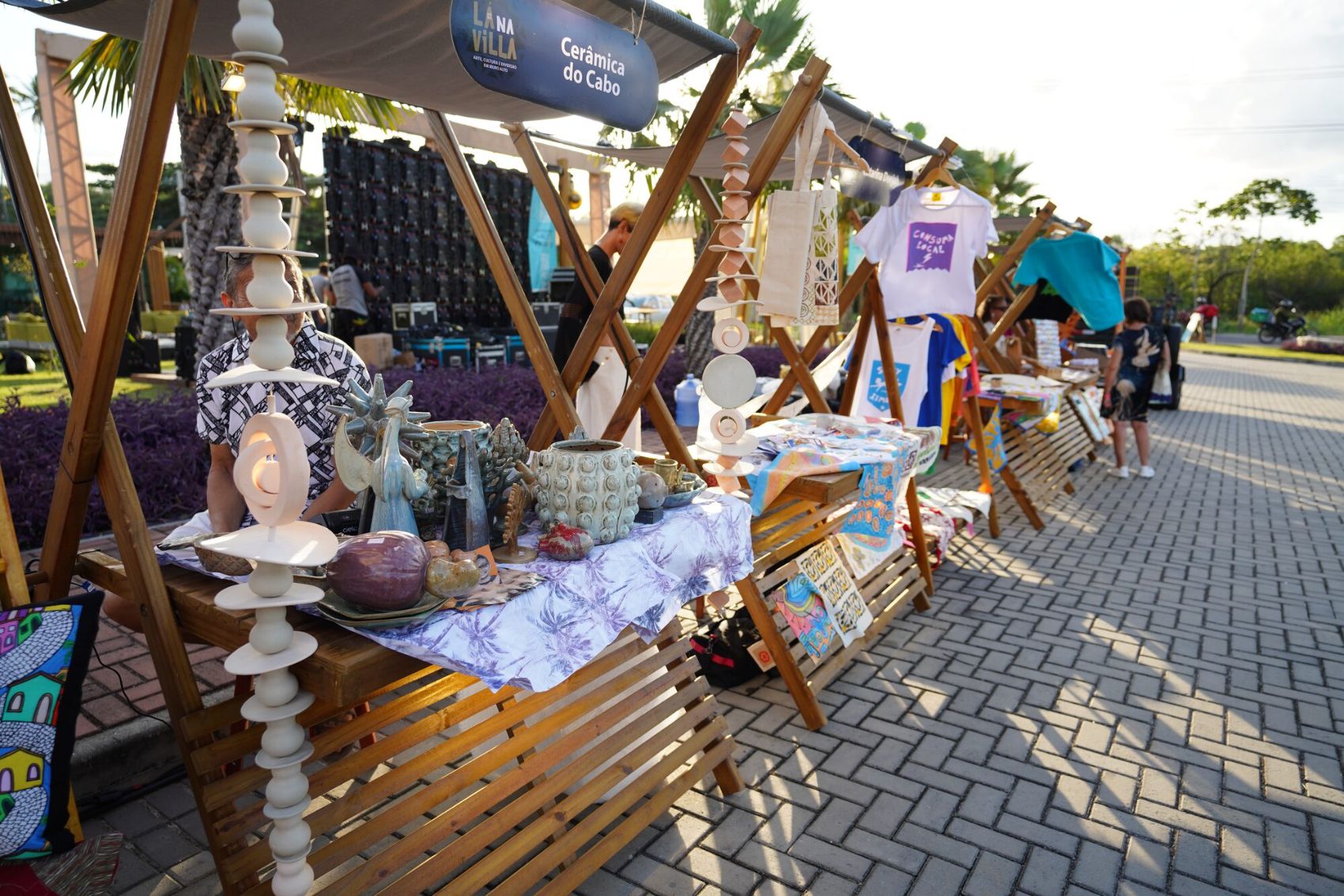 Feira criativa da Villa Muro Alto ao entardecer: barracas de madeira com cerâmica do Cabo, camisetas e peças autorais de artesãos da região.