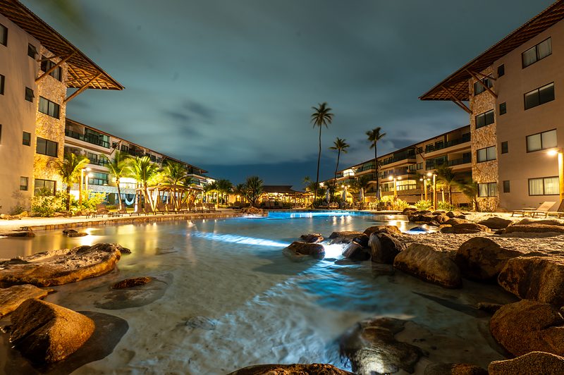 Piscina de borda de areia iluminada à noite entre os blocos residenciais do resort.