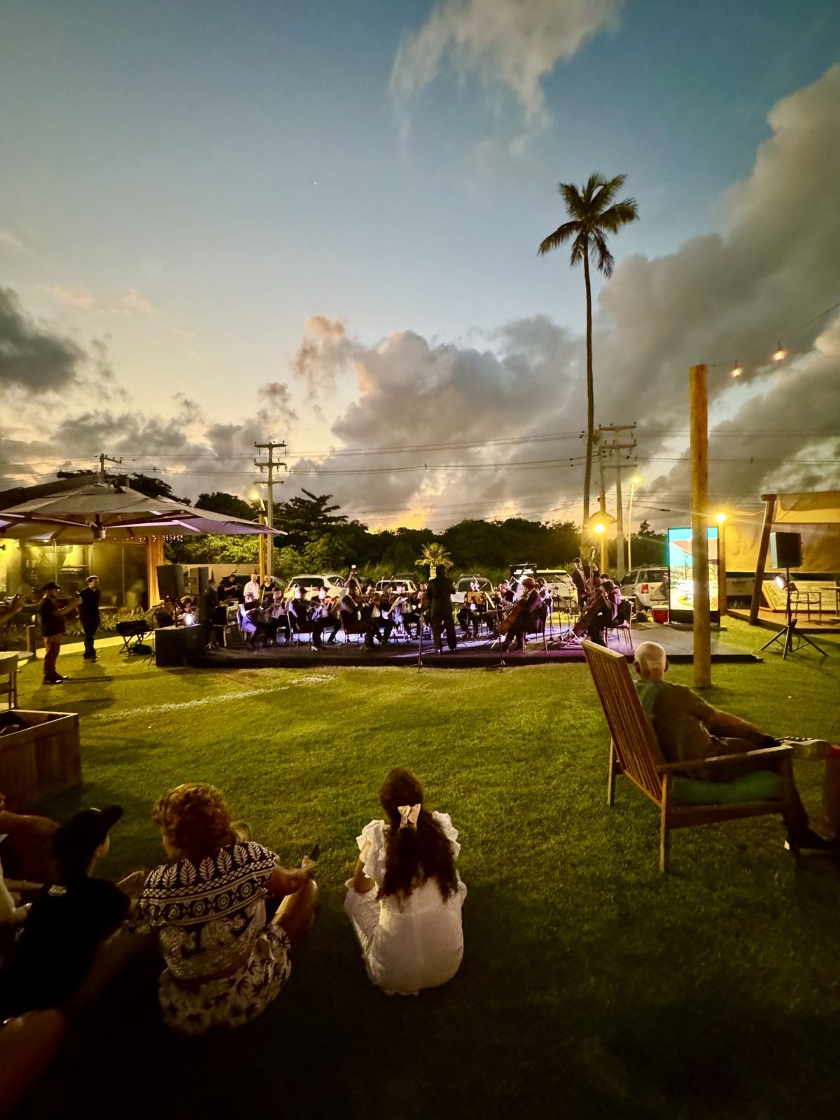 Concerto de orquestra ao entardecer no gramado da Villa, com público sentado na grama e nas cadeiras de madeira.