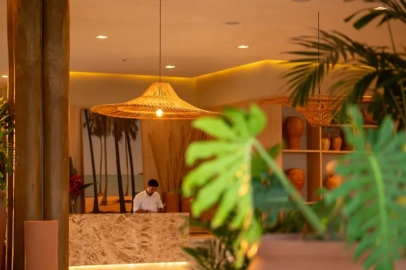 Recepção do resort com pendentes de fibra natural, bancada em pedra e vegetação.